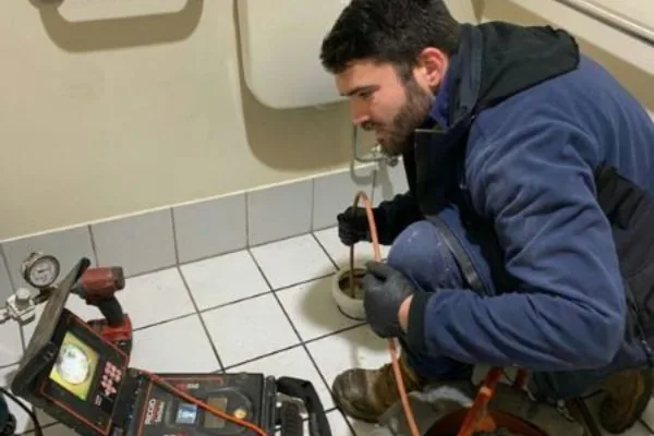 Plumber feeding a drain camera into a floor waste with the camera screen beside him