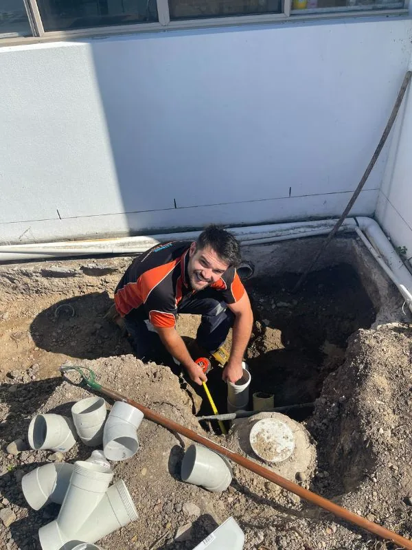 Linken crouching in an excavated pit, measuring a sewer junction with new PVC fittings laid out